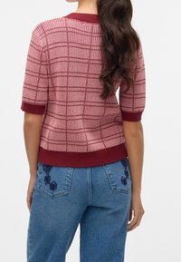 Femme aux longs cheveux foncés portant un pull à manches courtes à carreaux rose et rouge et un jean bleu avec une broderie florale sur les poches arrière.