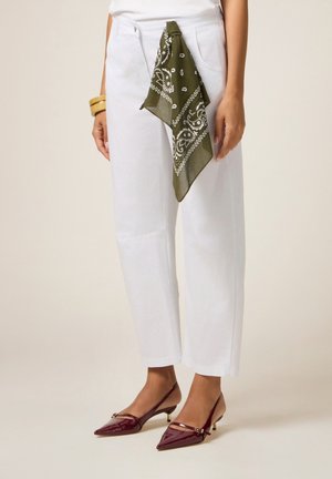 Kobieta ubrana w białe spodnie z oliwkowo-zielonym paisley bandaną w kieszeni, złote bransoletki i bordowe, szpiczaste buty na obcasie typu slingback.