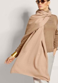 Hellbeige Schal, der um die Schultern gewickelt ist und aus weichem Stoff besteht. Über einem braunen Rollkragenpullover getragen, mit einem metallisch goldenen Armreif.