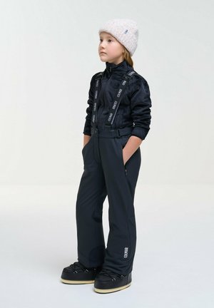 Un enfant pose dans une tenue de ski noire avec des bretelles et des bottes. Il porte un bonnet blanc en tricot et garde les mains dans les poches.