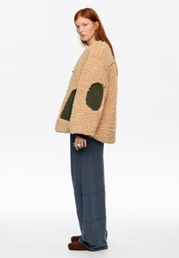 Joven mujer con largo cabello rojo, vestida con una chaqueta beige de piel con parches verdes, pantalones sueltos azules y zapatos marrones, posando de perfil.