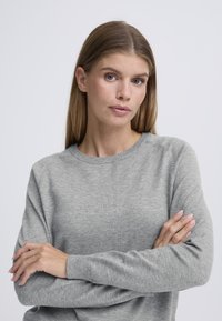 Pull gris clair tricoté avec un col rond, tissu texturé et poignets côtelés. Le modèle a les bras croisés devant lui.