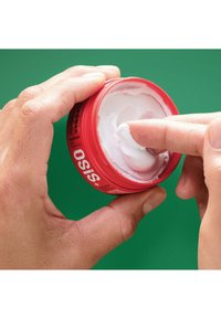 Röd rund behållare märkt "OSIS+" med vit hårkräm inuti. Ett fingertopp tar upp krämen. Grön bakgrund.