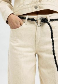 Beige denimbroek met een gestructureerde stof, voorzien van een zwart gevlochten riem en een metalen zilveren lus, met een zijzak die gedeeltelijk zichtbaar is.