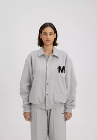 Grijze button-up sweatshirt met kraag, oversized pasvorm en zwart geborduurd "M" logo op de linkerkant van de borst. Zachte textuur en geribbelde manchetten.