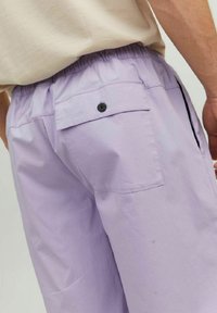 Lilac cotton shorts with an elastic waistband, a rear pocket secured by a black button, and a smooth, lightweight texture.