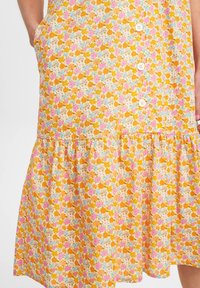 Vestido floral com fundo amarelo, apresentando flores rosa, laranja e azuis. A parte inferior é em camadas com babados, e tem três botões brancos na frente.