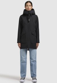 Parka imperméable noire avec capuche réglable, deux poches frontales et fermeture éclair. Associée à un jean bleu clair et des baskets noires.