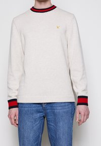 Pull en laine crème avec un col rond, des poignets rayés noirs et rouges, et un petit logo jaune sur la poitrine. Conception ajustée.