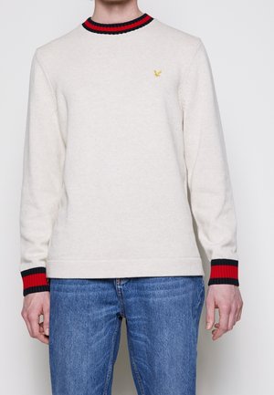 Pull en laine crème avec un col rond, des poignets rayés noirs et rouges, et un petit logo jaune sur la poitrine. Conception ajustée.