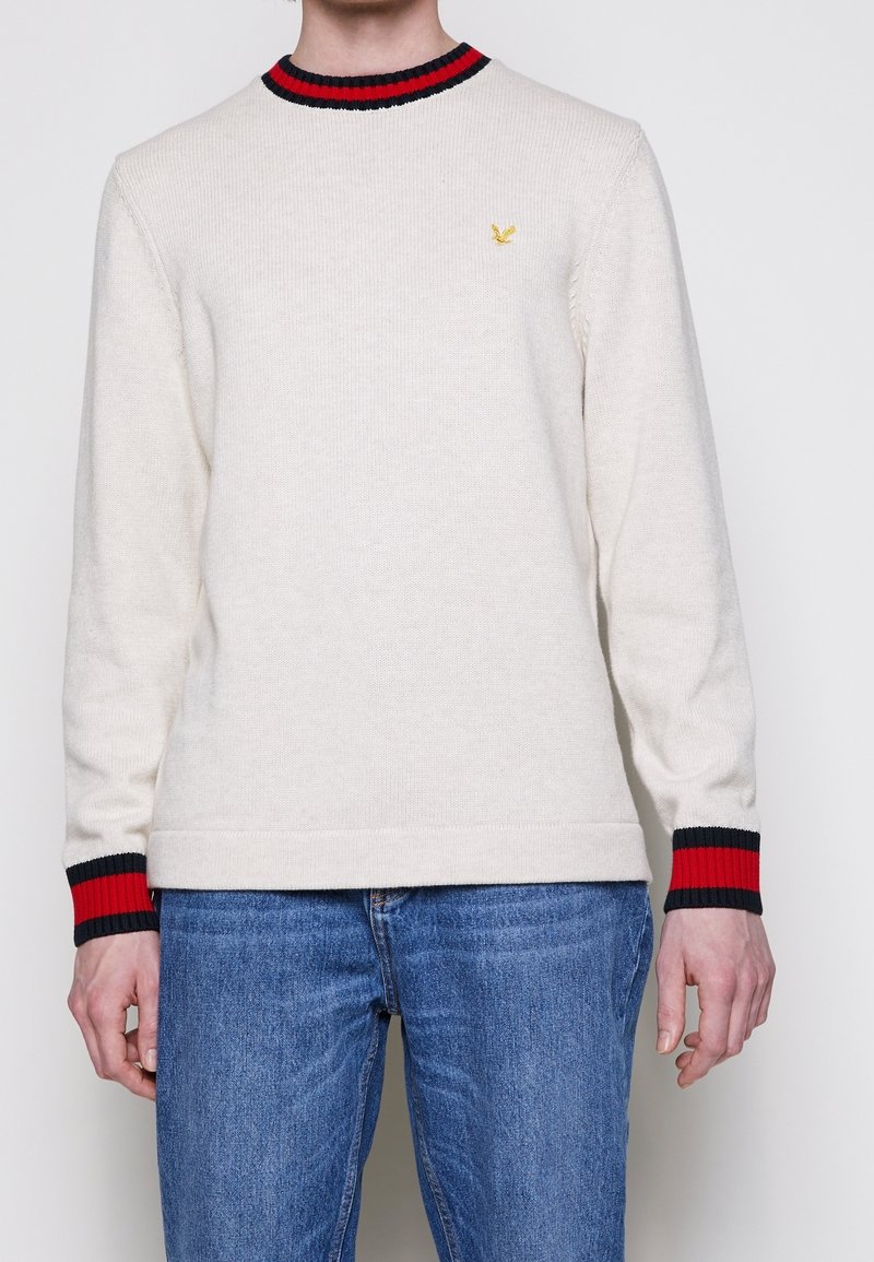 Pull en laine crème avec un col rond, des poignets rayés noirs et rouges, et un petit logo jaune sur la poitrine. Conception ajustée.