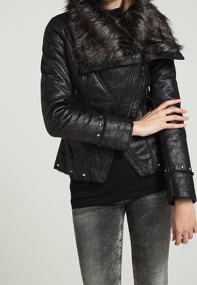 Veste noire en cuir synthétique avec col en fourrure, fermeture éclair à l'avant, accents cloutés et coupe ajustée, portée avec un jean gris déchiré.