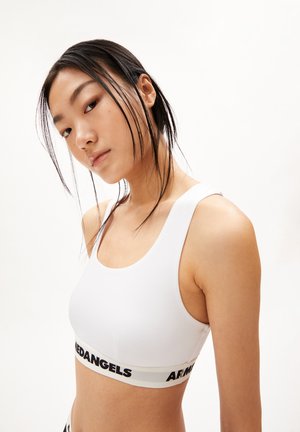 White sports bra with a scoop neckline, made of soft fabric. Features a black logo band at the bottom. Smooth texture and fitted design.