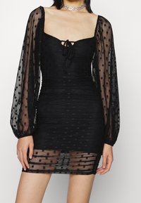 Musta mini-mekko, jossa on läpikuultavat pilkulliset hihat, rypytetty pinta, neliönmuotoinen pääntie ja sidontayksityiskohta edessä.