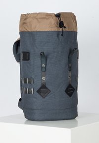 Sac à dos en textile gris avec un dessus brun, équipé de bretelles, de fermetures à pression et d'un cordon de serrage. Tissu texturé avec plusieurs accents et pièces métalliques.