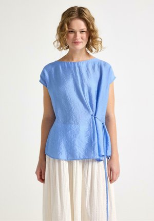 Giovane donna con capelli biondi ricci che indossa una blusa azzurro chiaro a maniche corte con fiocco laterale e una gonna bianca strutturata, in piedi davanti a uno sfondo semplice.