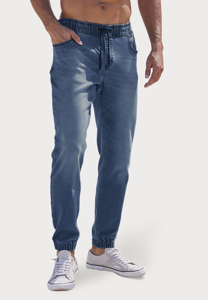 Homme portant un pantalon jogger en jean bleu avec des poignets élastiques et des baskets blanches, debout avec une main dans la poche devant un fond uni.
