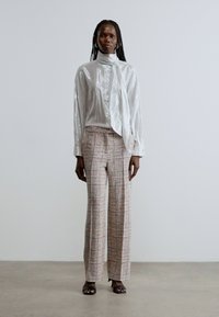Silvery, semi-transparent blouse with a tie collar; plaid, wide-leg trousers in pastel colors; model wearing black open-toe sandals.