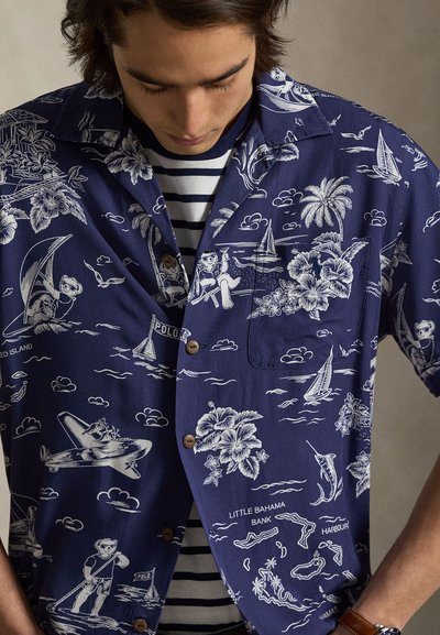 Młody mężczyzna ubrany w granatową hawajską koszulę z białymi tropikalnymi i żeglarskimi wzorami, noszoną na czarno-białej koszulce w paski.