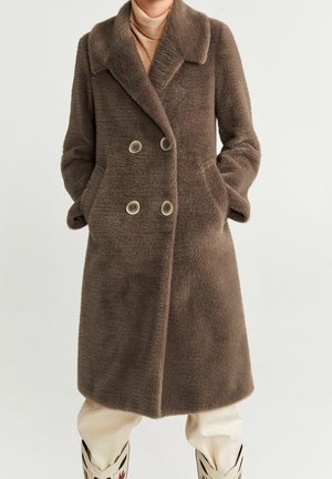 Femme portant un manteau marron mi-long à double boutonnage en tissu duveteux avec des poches latérales, un col roulé beige, un pantalon crème et des bottes noires et blanches.