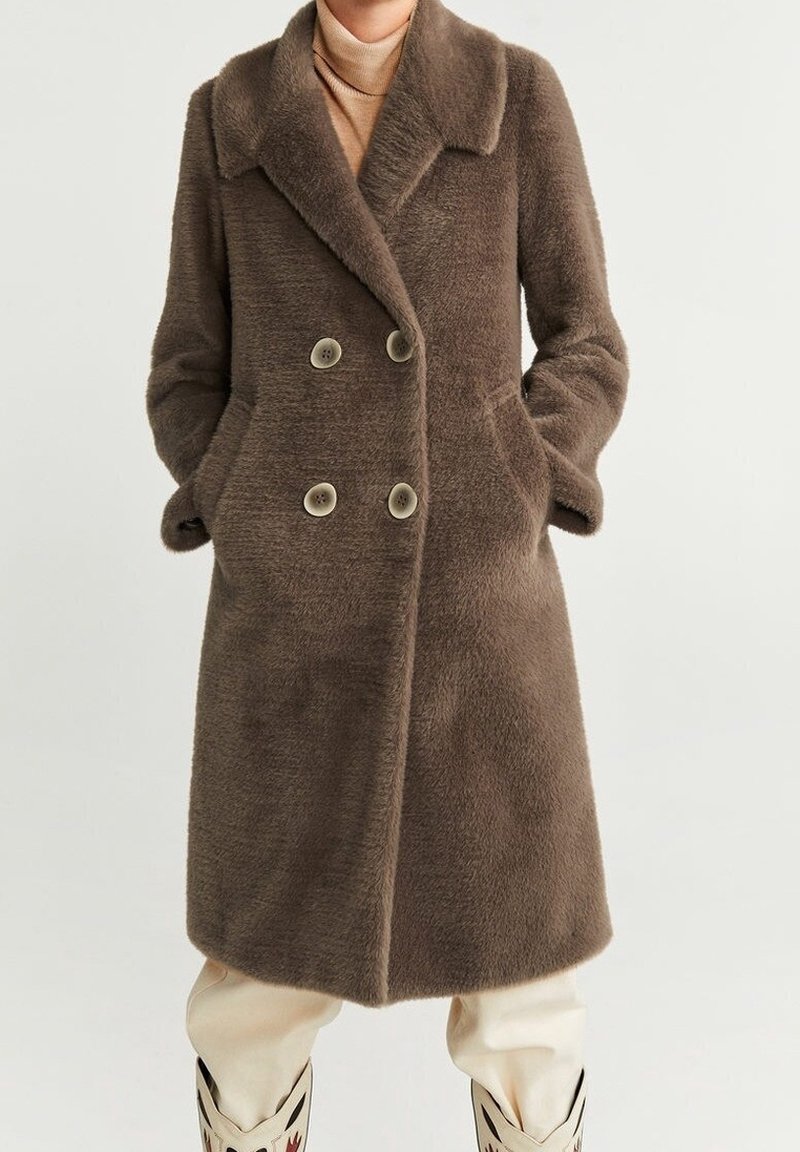 Femme portant un manteau marron mi-long à double boutonnage en tissu duveteux avec des poches latérales, un col roulé beige, un pantalon crème et des bottes noires et blanches.