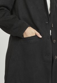 Cappotto in misto lana nero con una vestibilità rilassata, caratterizzato da una tasca sul lato sinistro, una chiusura a bottoni sul davanti e un tessuto leggermente testurizzato.