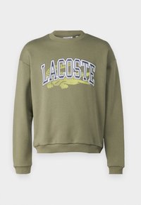 Sudadera verde oliva con cuello y puños acanalados. Presenta el texto "LACOSTE" en blanco y amarillo con un gráfico de cocodrilo verde debajo.