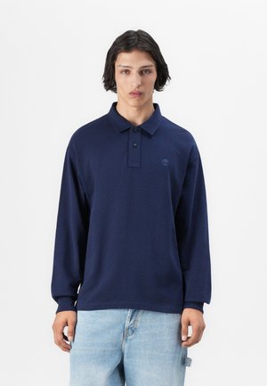 Navy long-sleeve polo shirt with a two-button placket and embroidered logo. Soft, smooth texture, relaxed fit, paired with light blue jeans.