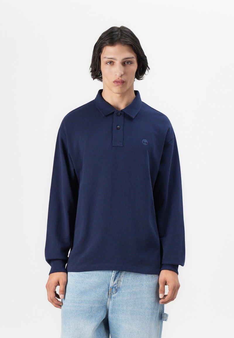 Polo de manga larga azul marino con una tapeta de dos botones y logo bordado. Textura suave y lisa, corte relajado, combinado con vaqueros de color azul claro.