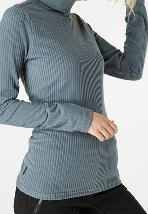 Personne portant un pull à col roulé côtelé bleu-gris ajusté et un pantalon noir, avec une main levée près de la tête.
