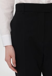 Czarne eleganckie spodnie z gładkiego materiału, z czystą talią i pasem na pasek oraz dopasowaną sylwetką. Widoczny mankiet białej koszuli.