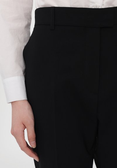 Juodi pritaikyti kelnės, pasižyminčios lygiu audiniu, švariu liemeniu su diržo kilpomis ir elegantiška siluetu. Matomas balto marškinio rankogalis.