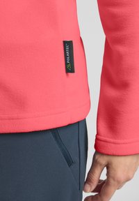 Pullover in pile di colore corallo brillante con finitura testurizzata. Presenta un'etichetta nera Polartec sull'orlo sinistro e polsini lisci. Abbinato a pantaloni scuri.