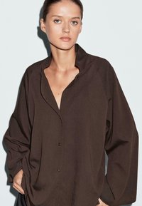 Bruine knoopshirt met een relaxte pasvorm, voorzien van een geribbelde textuur, een lage halslijn en lange mouwen. Subtiele details in de stof.