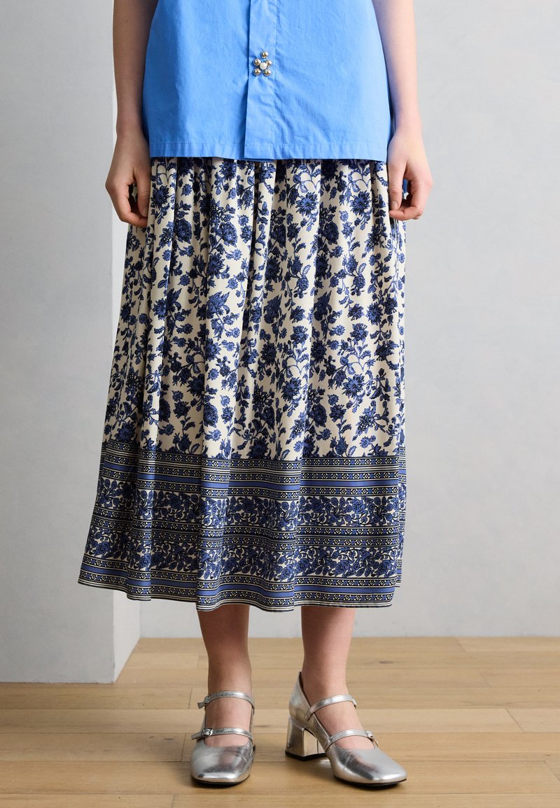 Άτομο που φοράει μπλε μπλούζα, μίντι φούστα με λευκό και μπλε λουλουδάτο σχέδιο και ασημί τακούνια Mary Jane, στέκεται σε ξύλινο πάτωμα.