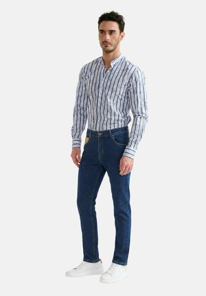 Uomo in piedi che indossa una camicia bianca e blu a righe maniche lunghe, jeans blu scuro e sneakers bianche su uno sfondo uniforme.