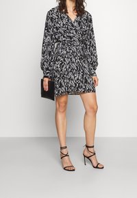 Svart och vit mönstrad omslagsklänning med långa ärmar och midjehögt bälte, kombinerad med svarta spetiga högklackade skor och en svart kuvertväska.