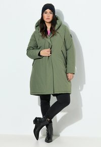 Parka di lunghezza media verde oliva con cappuccio, zip frontale e tasche laterali. Abbinata a jeans skinny neri e stivaletti.