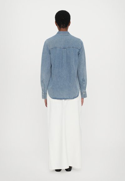Denim shirt in light blue, featuring a collar, long sleeves, and button cuffs. Worn over a white maxi skirt, with a smooth texture.