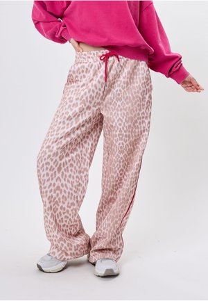 Kobieta nosząca luźne, jasnoróżowe spodnie w panterkę z czerwonym sznurkiem i białe trampki, zestawione z jasnoróżową bluzą.