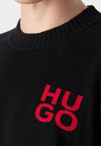 Pull en maille noire avec un col côtelé. Présente un patch logo rouge proéminent avec les lettres audacieuses "HUGO" sur la poitrine. Texture douce.