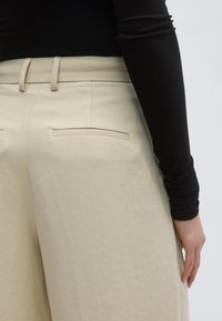 Μπεζ παντελόνι φαρδιάς γραμμής με δύο πίσω τσέπες, θηλιές για ζώνη και λεία υφή, συνδυασμένο με στενή μαύρη μπλούζα.