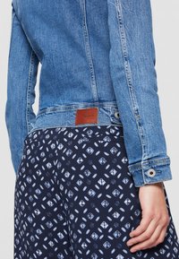 Chaqueta de mezclilla azul con puños abotonados sobre falda o pantalones con estampado azul marino, mostrando la vista trasera desde la cintura hasta la mitad del muslo con la mano visible.