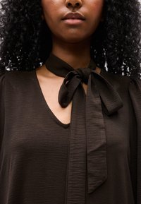 Γυναίκα με σγουρά μαλλιά που φοράει μια σκούρα καφέ μπλούζα με V-λαιμόκοψη και δεμένο φιόγκο στο γιακά.