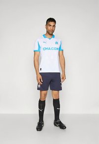 Camisola de futebol branca com detalhes em azul claro, logótipo da CMA CGM, combinada com calções azuis escuros, meias pretas e chuteiras. Design leve e desportivo.