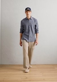 Chemise à rayures bleues, pantalon beige clair et chaussures beiges. Modèle portant une casquette marine ; arrière-plan avec un mur uni et un sol en bois.