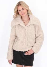 Giacca in pile beige con una superficie testurizzata e soffice, ampio colletto a punta e maniche lunghe, chiusura a bottoni frontale, indossata sopra una gonna scura.