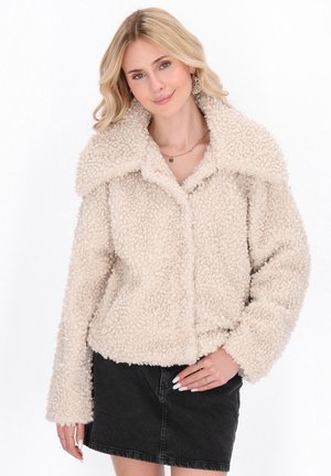 Beige fleecejas met een gestructureerd, pluizig oppervlak, grote geknipte kraag en lange mouwen, knoopsluiting aan de voorkant, gedragen over een donkere rok.