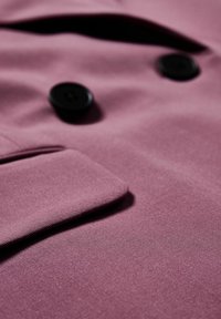 Gros plan sur un tissu mauve avec deux boutons noirs et un rabat de poche replié, mettant en valeur la texture et les détails du vêtement.