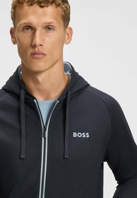Marineblauwe zip-up hoodie met koordjes, met een lichtblauwe binnenvoering en een witte "BOSS" logo op de borst. Glad stofweefsel.